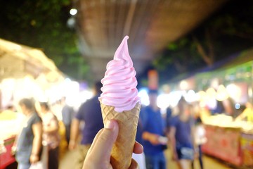A female hand holding a soft cone of strawberry yogurt ice cream with blurred many people walking around the night market area 