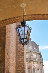 Fototapeta na wymiar An ornate cast iron lamp with frosted glass hangs from the bottom of a bridge with the tower of Toledo’s cathedral coming into view