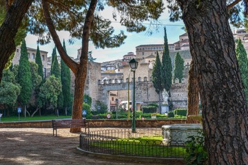 The city gate of Toledo, Spain seen through trees in an adjacent park. The battlements of the gate and defensive wall gives way to ever higher buildings as the city on the hill rises steadily upwards.