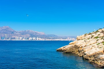 Light yellow rocks rise from the blue waters of the Mediterranean at an angle, with the tourist city of Benidorm, Spain in the background.