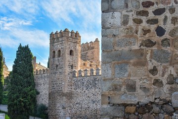 The defensive stone walls of Toledo, Spain, showing the battlements on top and a guard tower coming into view from behind an even closer wall, showing the detail of the ancient stone and mortar struct