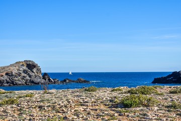 The dry semi-desert of southern Spain meets the blue water of the mediterranean with rocks rising above the water and a small sailboat sailing, along the coast.
