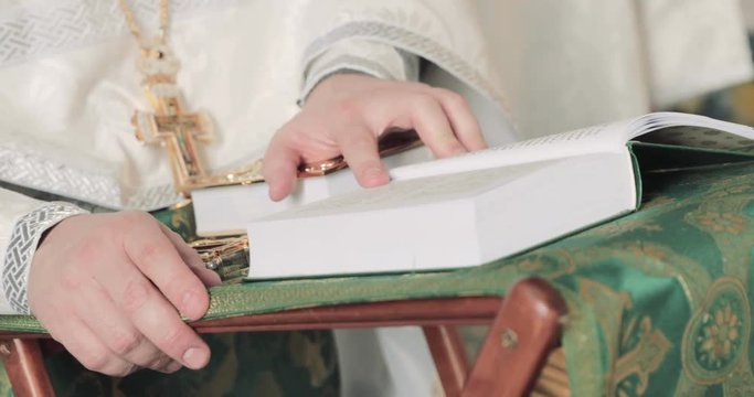 Hands of the priest leaf through pages of the religious book, is dressed in a cassock, a gold cross on a chain, a table-top with the beautiful embroidered fabric, the bible book with a gilded cover
