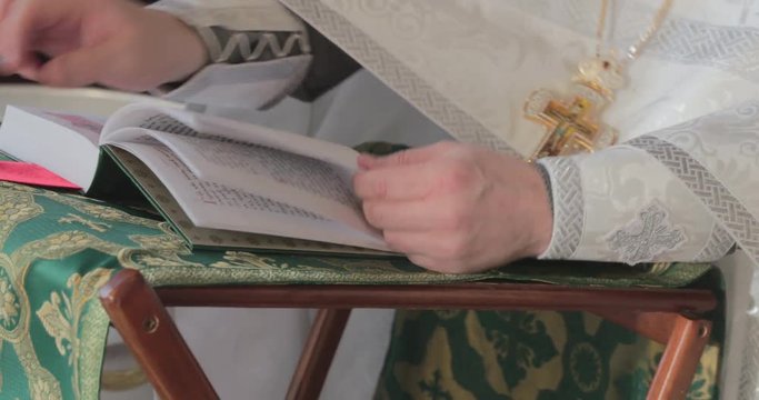 Hands of the priest leaf through pages of the religious book, is dressed in a cassock, a gold cross on a chain, a table-top with the beautiful embroidered fabric, the bible book with a gilded cover