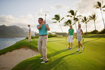 Man and two women watch a male golfer take a shot over a beach sand trap.