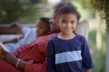 Portrait of a young boy standing beside his mature grandmother lying on a couch reading with his brother.