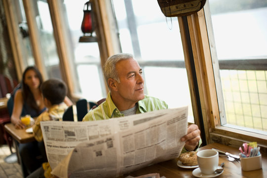 Mature man looking out a window while holding a newspaper inside a cafe.