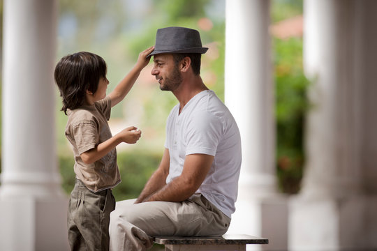 Thirtysomething man sits as his young son plays with his trilby hat.