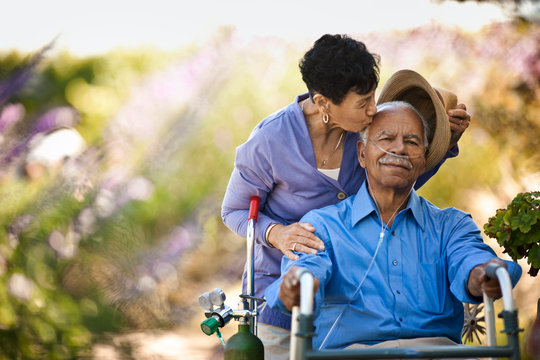 Happy senior man in a wheelchair being kissed on the head by his wife in a garden.