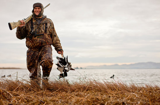 Satisfied duck hunter wearing camouflage clothing carrying two dead ducks and shotgun as he walks along edge of a lake.