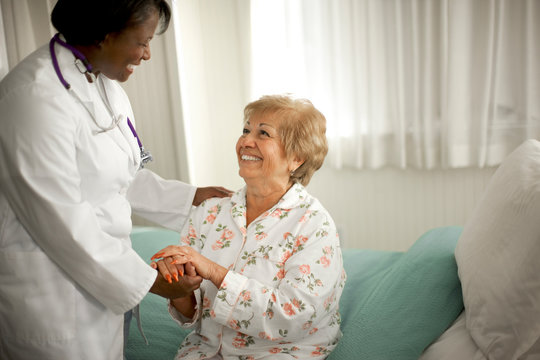 Smiling senior woman holding the hand of a female doctor as she sits on the edge of a bed inside a room.