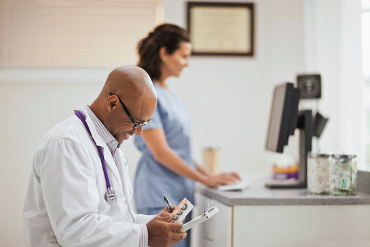 Smiling male doctor taking notes on a clipboard as he holding a box of medication while in his office with a nurse.
