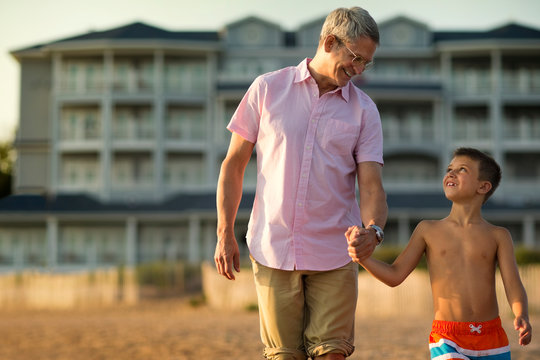 Father smiling and talking with his young son as they hold hands and walk on the beach at sunset.