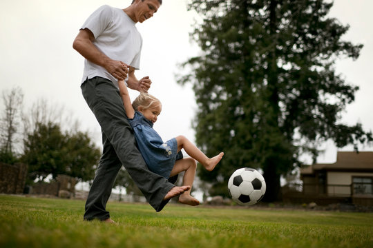 Smiling father teaches his little girl how to kick a soccer ball on the lawn.