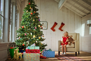 Young girl sitting on an armchair with her teddy bear, next to a Christmas tree.