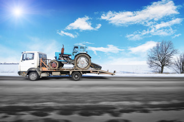 Tow truck driving fast with old tractor on the winter countryside road against blue sky with sun