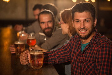 Handsome man in checked shirt holding glass of beer while sitting in bar. Bearded man smiling and looking at camera. Background of people talking and resting in beer pub.