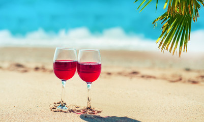 Two glasses with red wine on the beach. Sea, sand and palm on background.