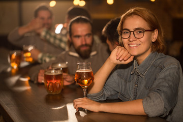 Adorable woman in spectacles posing in beer pub full of clients. Young woman smiling, looking at camera and holding hand near chin. Female client holding glass of delicious beer.