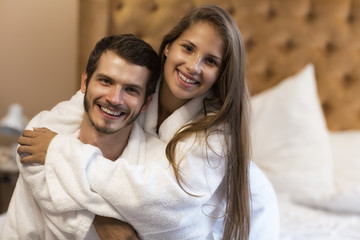 Fototapeta na wymiar Young couple in love posing in comfortable suite in hotel. Pretty woman with chestnut hair hugging her husband. Handsome bearded man and gorgeous woman looking at camera and smiling.
