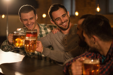 Young and cheerful men sitting in pub and having fun. Positive male clients holding glasses of tasty beer and clinking. Handsome men smiling, resting and communicating.