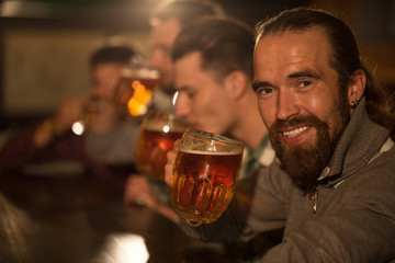 Close up of happy handsome bearded man showing glass of beer in hand. Male client of beer pub smiling and looking at camera. People drinking beer and resting in bar on background.