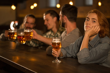 People sitting in beer pub and hanging out. Sad woman with ginger hair being bored while men having fun and clinking beer glasses. Beautiful young woman looking at camera.