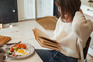 young girl is reading an old book at breakfast. Close-up hands and dinner table setting.