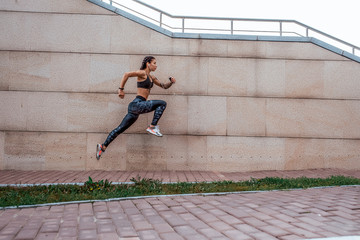 Beautiful young girl tattoos summer city runs jump in hand smartphone listens music in headphones, sportswear leggings bra. Free space text. Morning exercise in fresh air on background concrete wall.