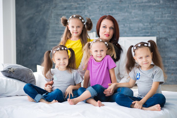 Obraz premium Large friendly family, many children: mom and four pretty cheerful girls triple twins sisters sitting on a bed against a gray brick wall