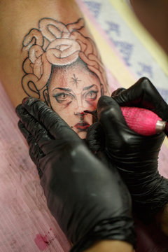 Close up of professional master drawing medusa gorgon tattoo on skin. Hands of unrecognizable tattooist in black gloves using black ing and tattoo machine for picture.