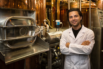 Fototapeta na wymiar Professional brewer in white coat standing near bronzed beer tank wearing in white coat. Handsome man smiling, posing and looking at camera. Background of beer factory and industrial equipment.