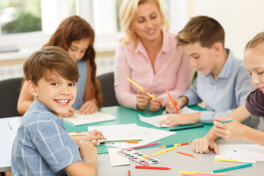 Pupils with teacher sitting at desk, enjoying painting with colorful pencil at school classroom and one boy smiling at camera. Girl explaining something to her classmate and woman helping children.