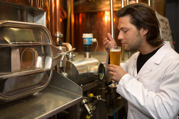Brewer wearing in white coat holding beer glass and smelling it. Handsome worker of brewery tasting beer with closed eyes. Specialist standing near steel machinery and equipment.