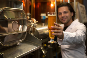 Close up of beer glass. Brewer wearing in white coat looking at glass of tasty beer and holding it in hand. Interior of brewery with professional steel machinery and industrial equipment.