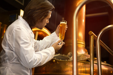 Specialist of brew industry holding glass of beer in hands and looking at it. Professional brewer checking and examining beverage on quality. Worker of brewery wearing in white coat.