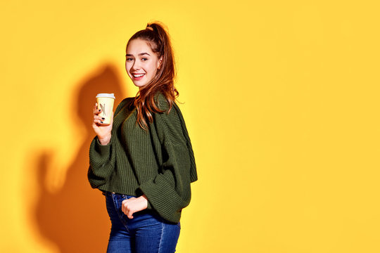 Image of pretty young woman smiling and holding takeaway coffee in paper cup isolated over yellow background