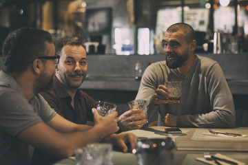 Three male friends sitting in bar and communicating. Men drinking alcohol beverages, whiskey or brandy. Cheerful men smiling, having fun and holding glasses with drinks.