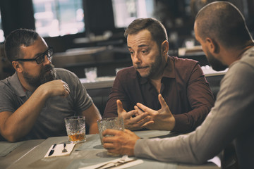 Friends talking and communicating while sitting and spending time together in bar. They drinking whiskey or scotch. Sad man in shirt having problems and men trying support their friend.