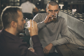 People sitting at bar counter and drinking alcoholic drinks. Male clients holding glasses of delicious whisky or scotch and tasting it. Interior of modern comfortable bar.