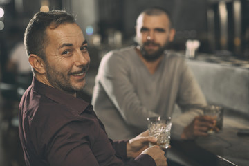Handsome man looking at camera, smiling and holding glass of delicious alcohol. Friend sitting at bar counter behind and posing. Men relaxing and spending time together in bar.