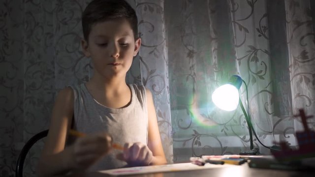 Little boy sitting at table and drawing with colored pencils in the light of a lamp
