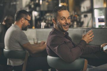 Positive client of bar sitting at counter in chair and looking back through shoulder at camera. Handsome man holding glass of strong alcohol and smiling. People sitting in bar behind.