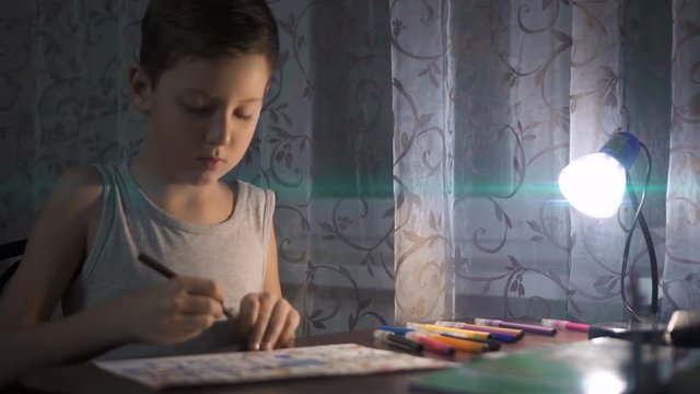 Little boy sitting at table and drawing with colored pencils in the light of a lamp