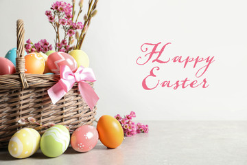 Wicker basket with colorful painted eggs on table and text Happy Easter against light background