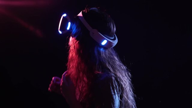 Young woman wearing VR headset making gesture on dark background illuminated neon red and blue lights