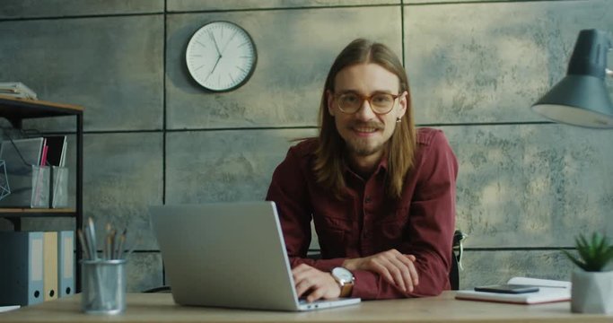 Portrait shot of the young Caucasian cheerful man in glasses working at the laptop computer in the office and then smiling to the camera.