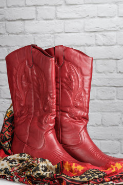red coyboy style cowgirl boots  with decorative stitching and a southwest pattern scarf in front of a white brick wall