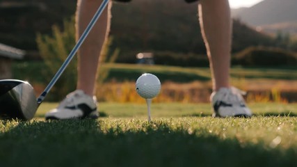 Close Up On Golf Ball Being Hit Off Tee With Sun In Background. Low angle, close-up shot showing a golf club that is swung, hitting the golf ball. The scene also shows a beautiful sunny background.