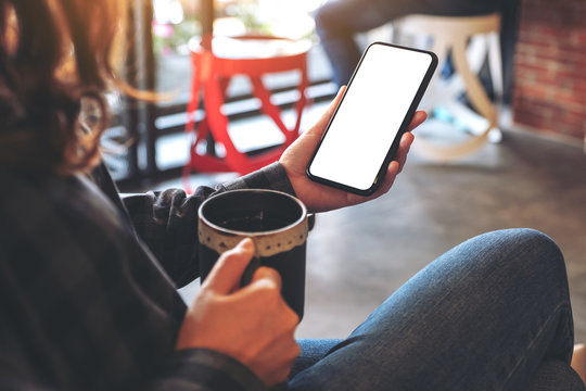 Mockup image of a hand holding black mobile phone with blank screen while drinking coffee in cafe